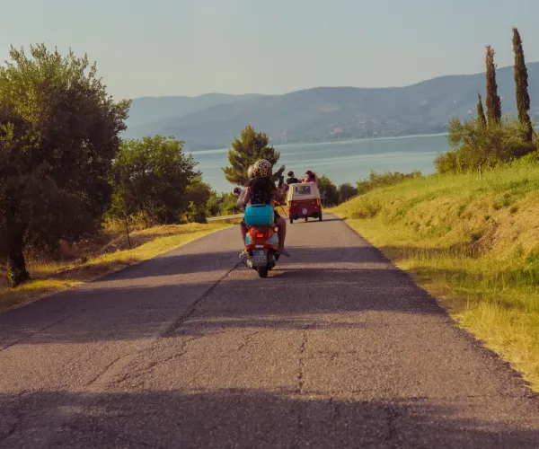 Person riding scooter on a country road toward a van, with trees and hills in the background.