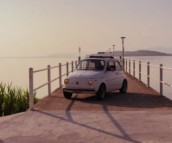 Vintage car on a pier at sunset, with calm water and soft pink sky.