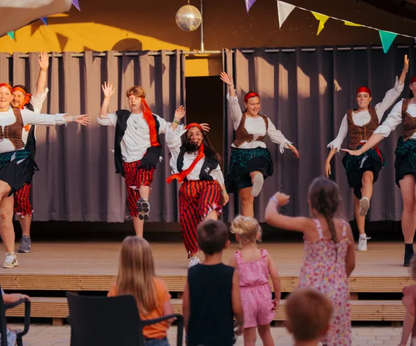 Dancers in costume perform on stage, watched by children in the audience.