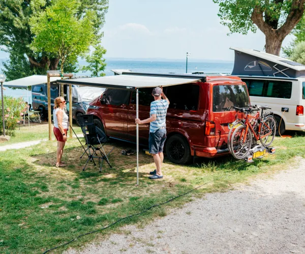 Camping scene with a red van near a lake, people setting up an awning, bikes on a rack.