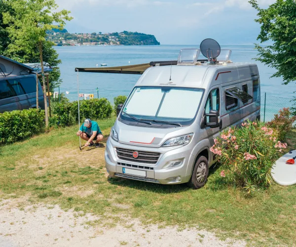 Camper van parked by a lake with a person sitting nearby, surfboard and greenery around.