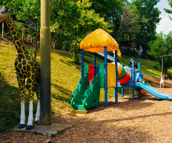Playground with colorful slide, giraffe statue, surrounded by trees.