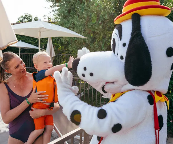 Child high-fives person in a Dalmatian mascot costume by poolside, held by smiling adult.