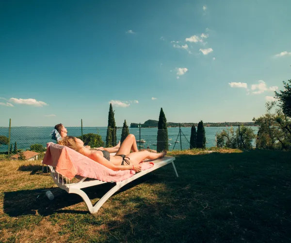 Couple sunbathing on chaise longue by a lakeside under a clear sky.