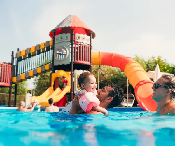 Family enjoying a sunny day at a pool with colorful slides in the background.