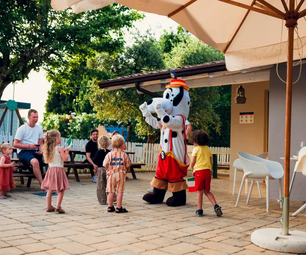 Children gather around a mascot in a sunny patio area.