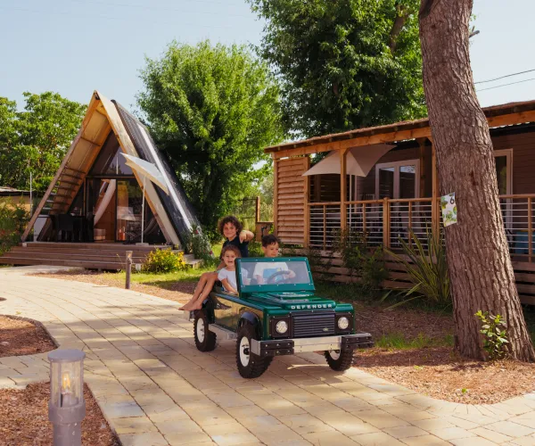 Two kids ride a mini green jeep on a sunny path by cabins.