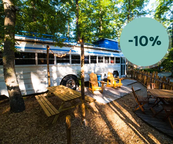 White bus converted into a campsite with chairs and tables, surrounded by trees.
