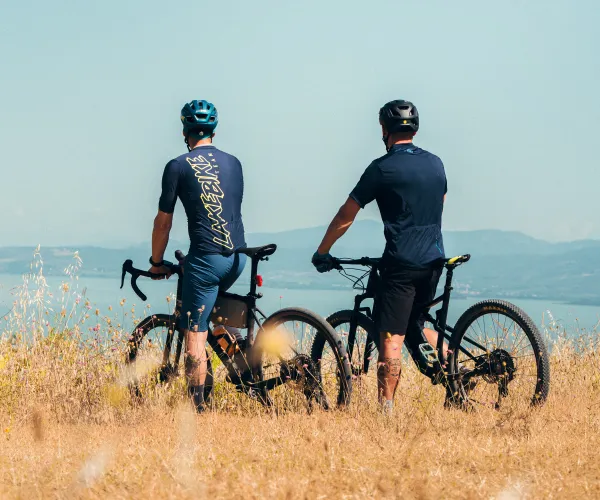 Two cyclists on a grassy hill overlooking a distant lake and mountains.